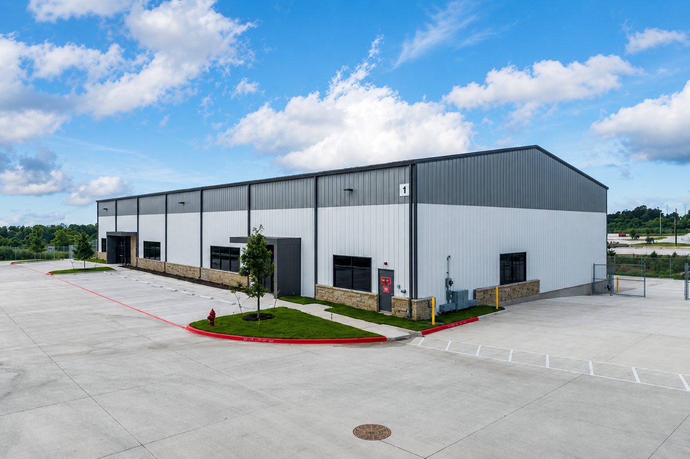Newly built single-storey industrial building with a grey and white metal facade, stone base, and striped parking court.